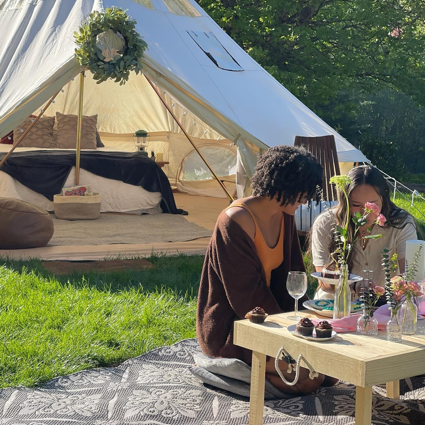 Two people sit at a low table with desserts and drinks on a patterned mat outside a furnished tent in a grassy, sunlit area.