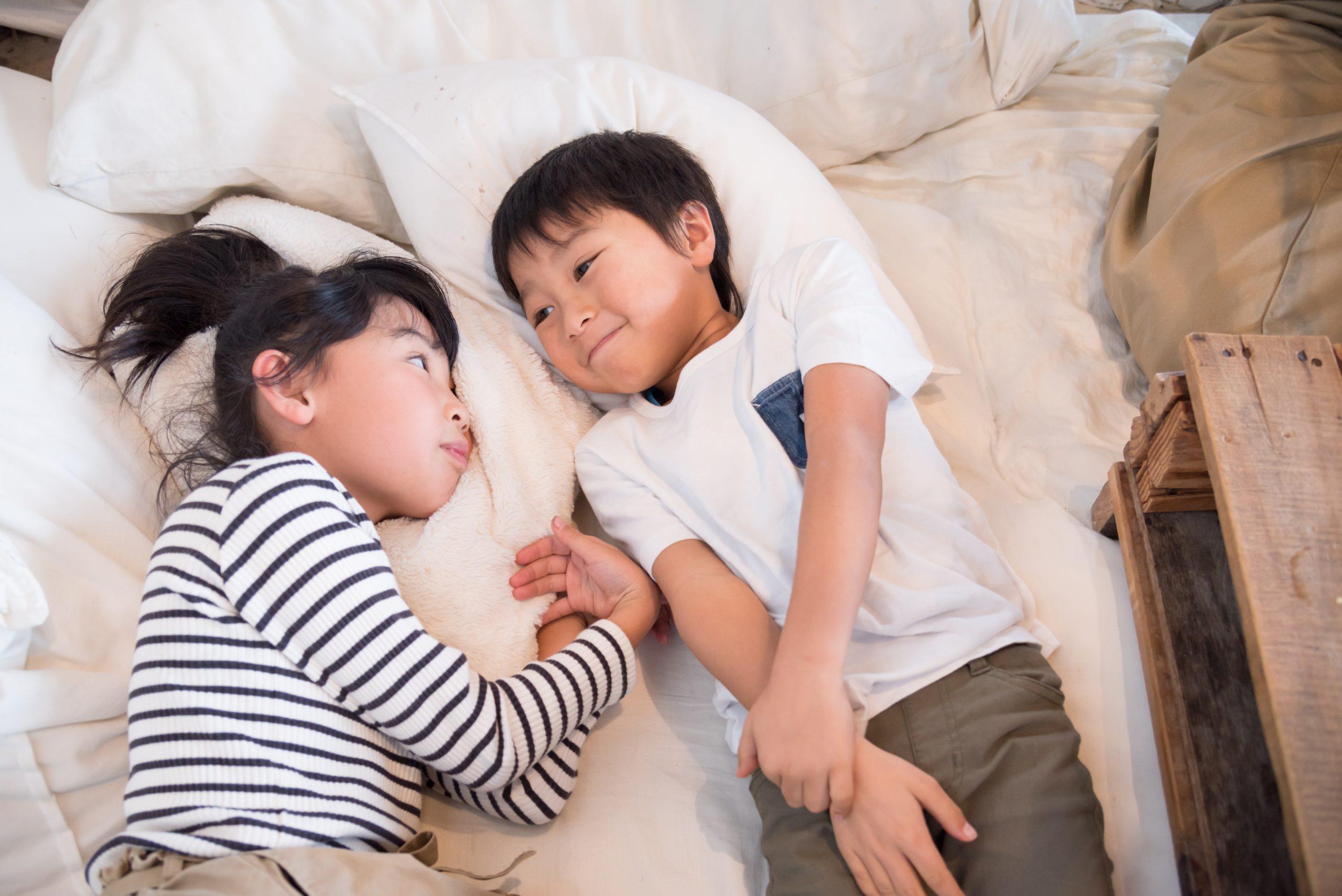 Two children lie next to each other on a bed, facing each other and appearing to have a conversation. They are surrounded by white bedding and a wooden surface is visible nearby.