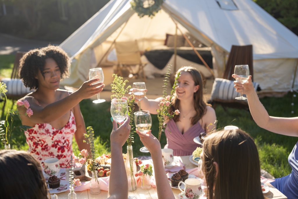 Four people sit at a decorated outdoor table in front of a tent, raising glasses in a toast. Sunlight illuminates the scene and floral arrangements are visible on the table.