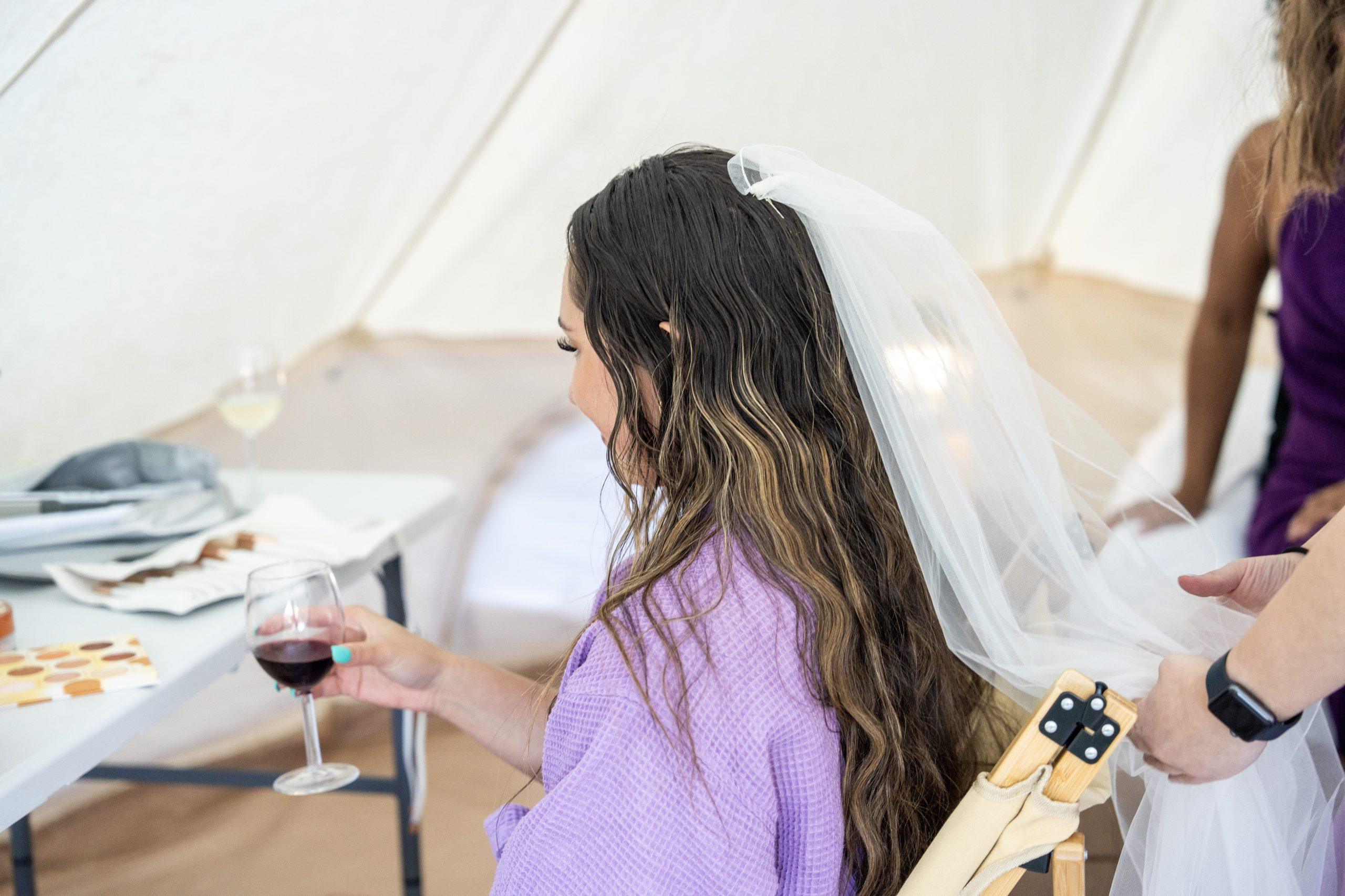 A woman in a purple robe and veil sits while someone arranges her hair; she holds a glass of red wine in one hand inside a tent.