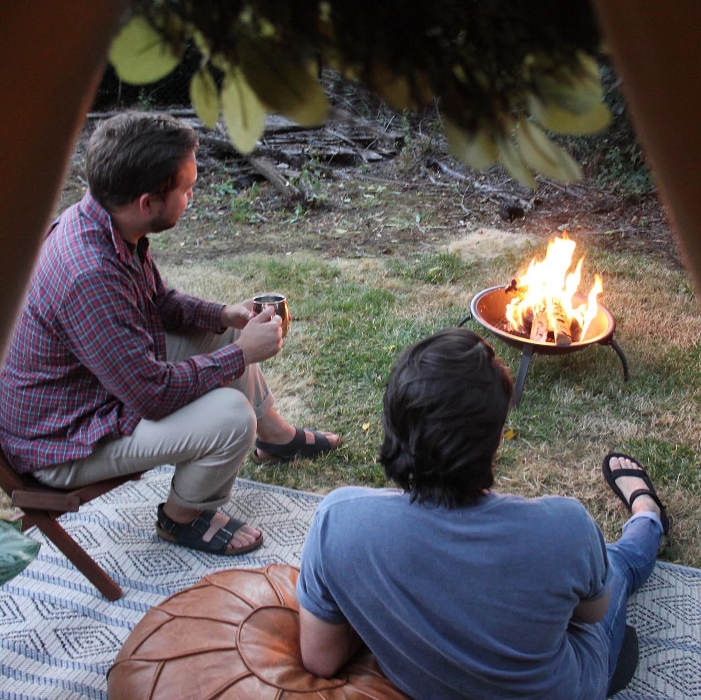 Two people sit on chairs by a small fire pit in a backyard, one holding a mug. The scene is viewed from behind, under a tent.