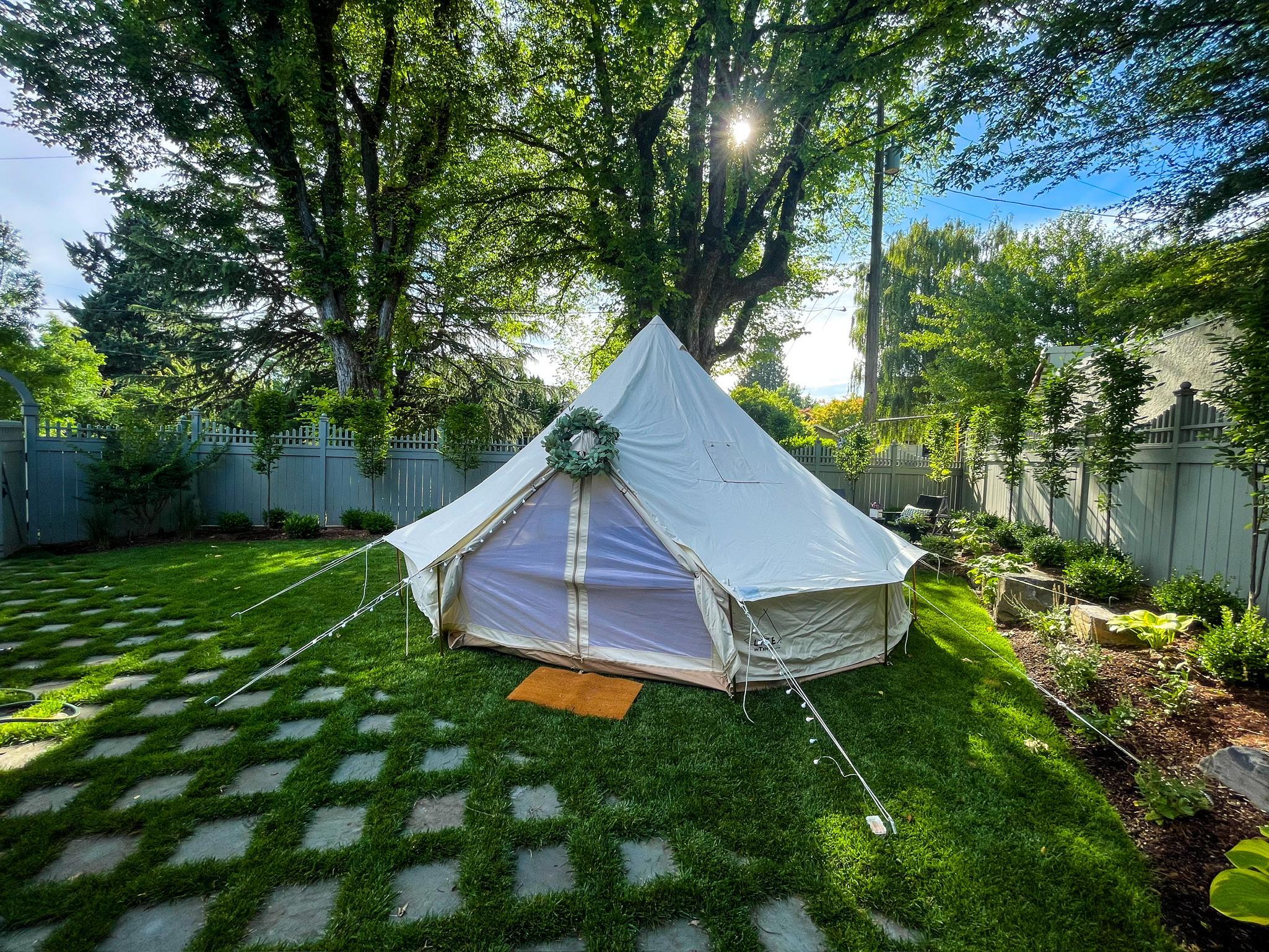 A white canvas tent with a floral wreath over the entrance is set up on a grassy lawn in a fenced backyard surrounded by trees and garden beds.