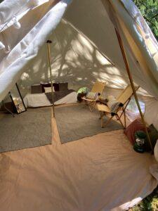 The inside of a canvas tent with a bed, two wooden chairs with cushions, rugs, a mirror, and a green lantern; sunlight filters through the fabric.