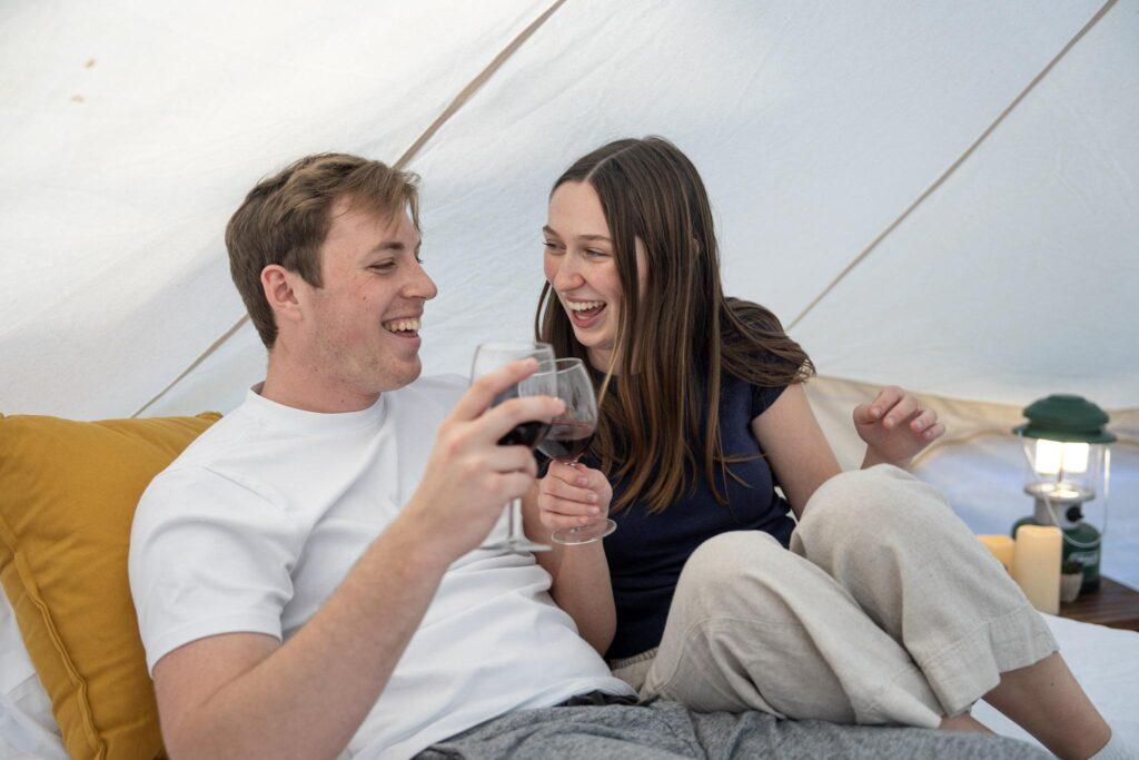 A man and a woman sit on a bed inside a tent, smiling and holding glasses of red wine. Pillows, a lantern, and candles are visible in the background.