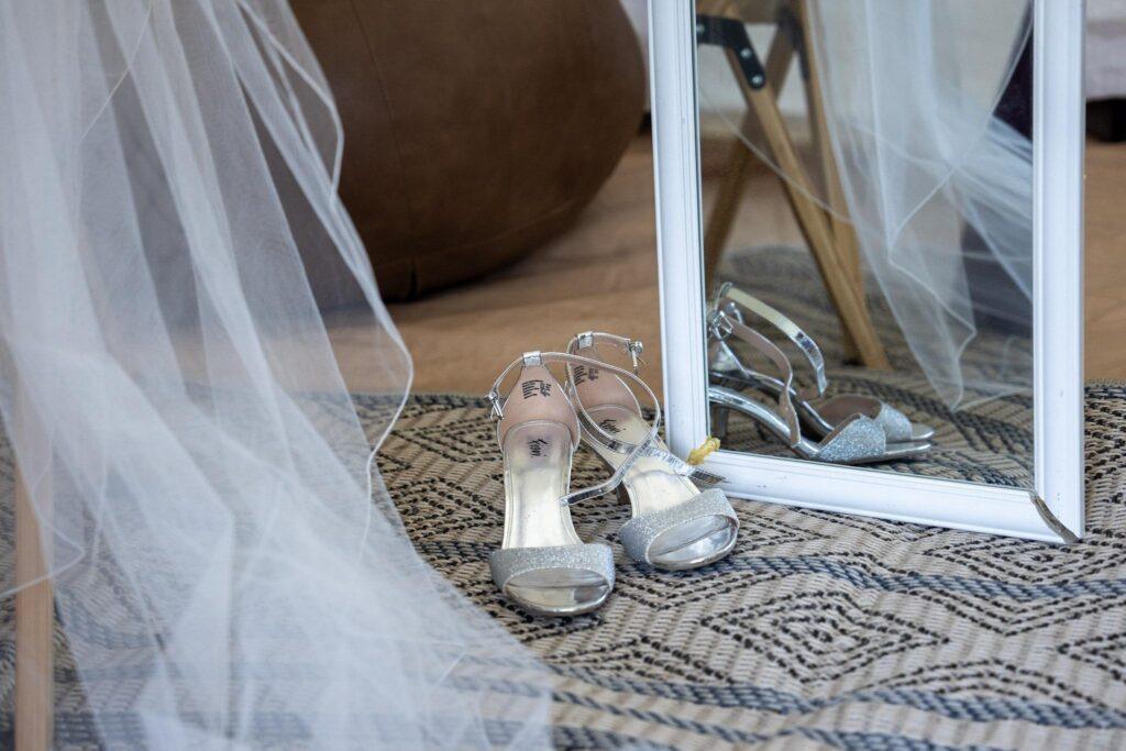 A pair of silver high-heeled shoes is placed on a patterned rug next to a large mirror, with a veil draped on the side.