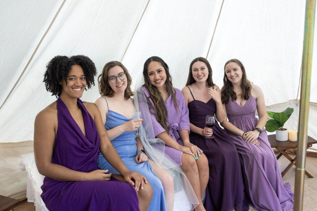 Five women in colorful dresses sit side by side on a bench, smiling at the camera inside a white tent, with drinks and a potted plant nearby.