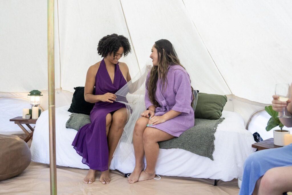 Two women sit on a bed inside a tent, talking and smiling. One holds a piece of fabric, and both wear purple dresses. Others are partially visible nearby.