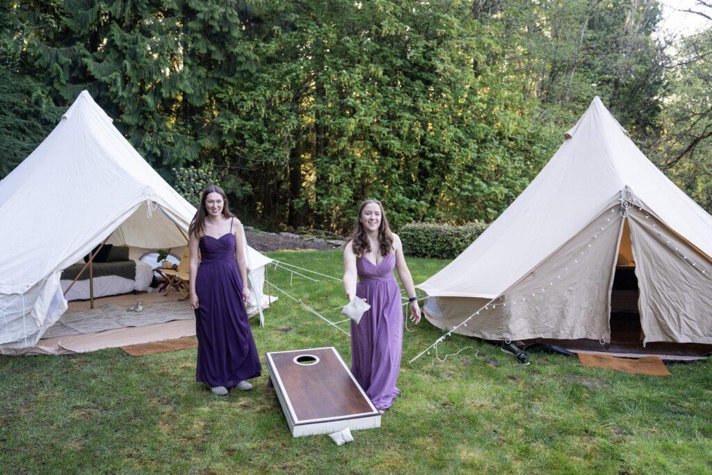 Two women in purple dresses stand on grass between two canvas tents, playing a game of cornhole in an outdoor setting surrounded by trees.