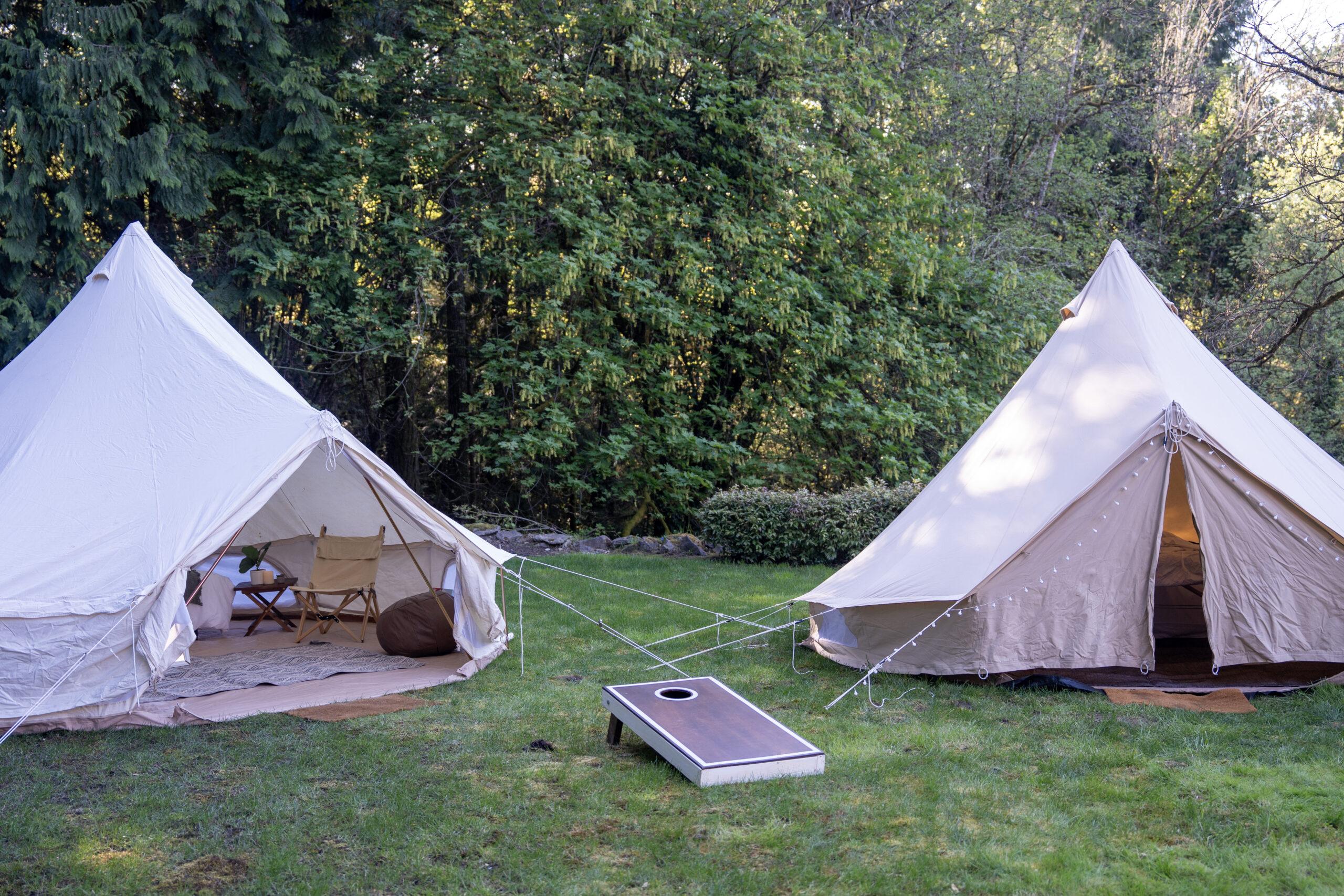 Two canvas tents are set up on grass with their entrances open, surrounded by trees. A cornhole game board is placed between the tents.